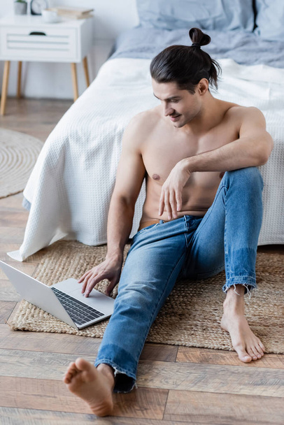 barefoot man with long hair smiling and using laptop on carpet near bed - Photo, Image