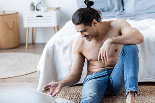 homme heureux avec les cheveux longs assis et en utilisant un ordinateur portable sur le tapis près du lit - Photo, image