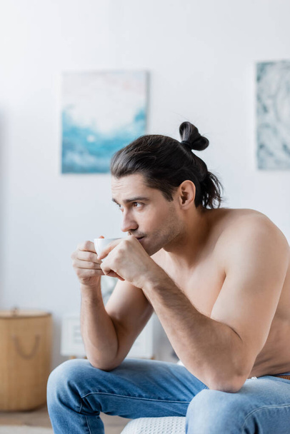 shirtless man with long hair holding cup of coffee while sitting on bed - Фото, зображення