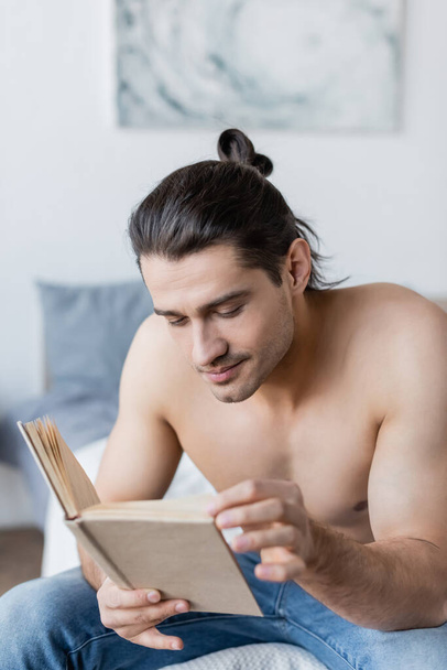 félmeztelen férfi hosszú haj olvasás könyv és ül az ágyon - Fotó, kép