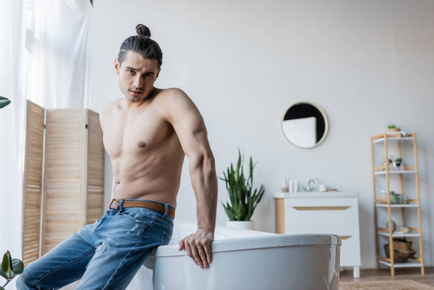 shirtless man with long hair sitting on bathtub in modern bathroom  - Foto, afbeelding