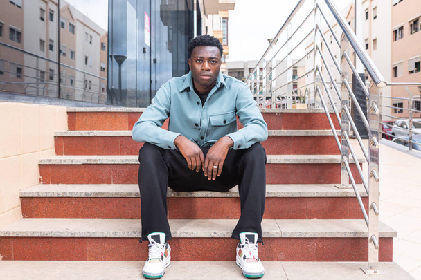 Full body of confident serious young African American male with curly dark hair in trendy outfit sitting on stairs of modern building and looking at camera in city - Photo, Image