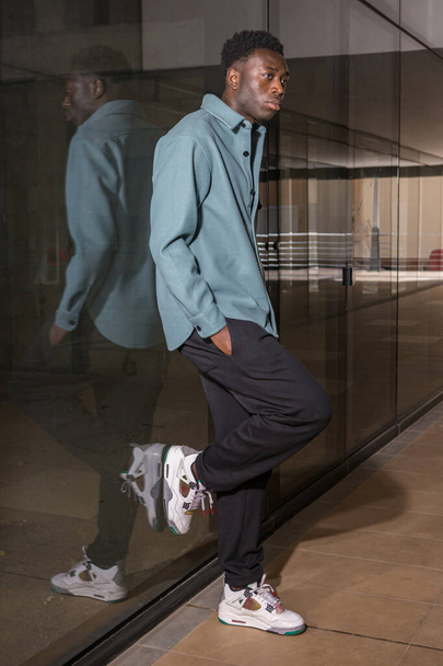 Seitenansicht Nachdenklicher junger afroamerikanischer Mann im trendigen Outfit und Turnschuhen lehnt an Glaswand im modernen Hausflur und schaut weg - Foto, Bild