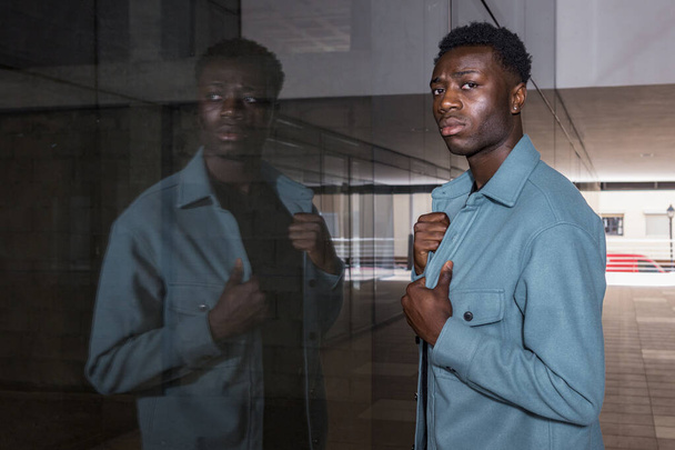 Widok z boku pewny siebie młody etniczny mężczyzna model z ciemnymi włosami stojący w pobliżu szklanej lustrzanej ściany nowoczesnego budynku i patrząc w aparat - Zdjęcie, obraz