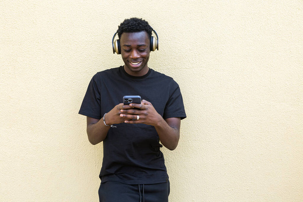 Joyful young confident African American guy in total black outfit and modern headphones listening to music and messaging on mobile phone standing near wall on street - Photo, Image
