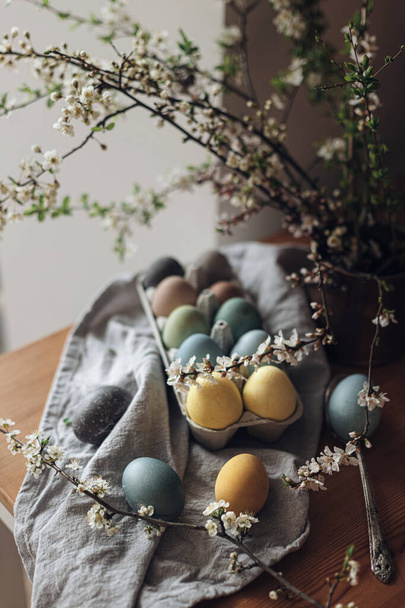 Easter eggs on rustic table with cherry blossoms. Happy Easter! Natural dyed colorful eggs in paper tray with linen napkin and spring flowers in rustic room. Countryside still life - Valokuva, kuva