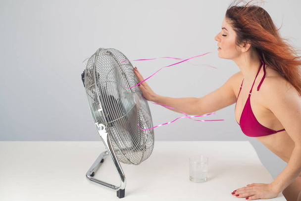 Red-haired smiling woman in a bikini drinks a cold drink and enjoys the blowing wind from an electric fan on a white background. Climate control on a hot summer day - Foto, afbeelding
