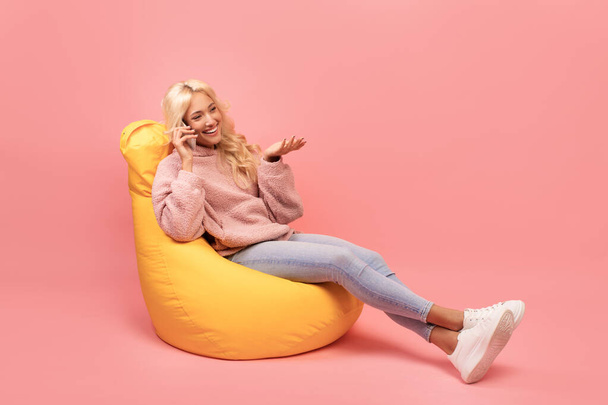 Mulher bonita falando no celular, fazendo chamada, sentado na cadeira beanbag contra fundo rosa, espaço de cópia - Foto, Imagem