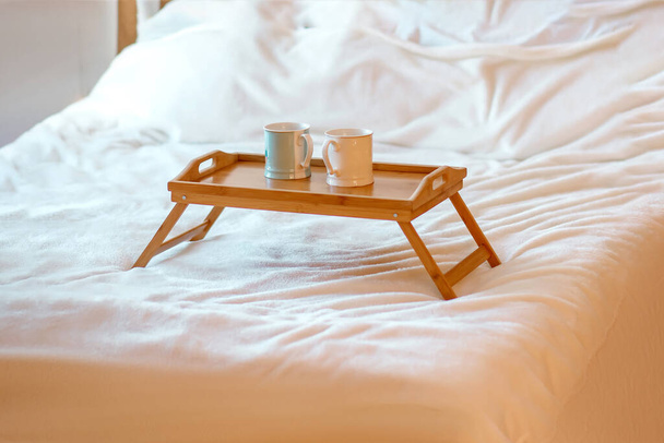 Two mugs with hot drinks on a wooden tray stand on a white bed. Cozy atmosphere or relaxation concept. Breakfast in bed in a cozy room - Foto, Imagem