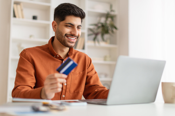 Χαμογελώντας νεαρός άνδρας χρησιμοποιώντας υπολογιστή και πιστωτική κάρτα στο σπίτι - Φωτογραφία, εικόνα