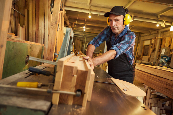 Ο πρεσβύτερος ξυλουργός με στολή εργάζεται σε ξυλουργική μηχανή στην ξυλουργική - Φωτογραφία, εικόνα
