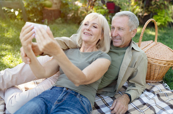 Glückliche Senioren telefonieren mit dem Smartphone beim Picknick und Ausruhen im Garten an warmen Frühlingstagen - Foto, Bild