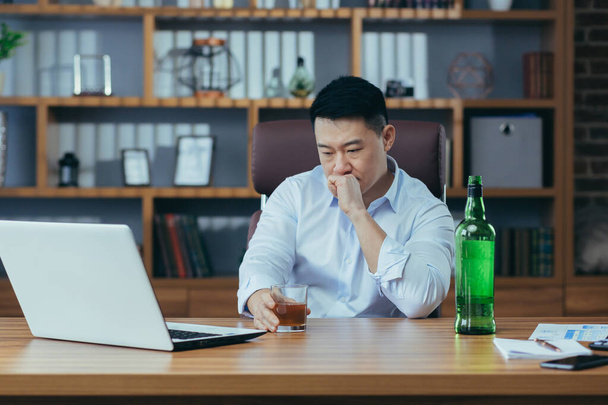 Ázsiai üzletember elbukik, kétségbeesett férfi iszik kemény italt, sokáig ül az asztalnál az irodában, depressziós és reménytelen. - Fotó, kép