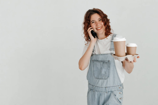 Fiatal nő overállal, kezében egy csésze forró kávéval, a fehér falon túl. A kapcsolattartás koncepciója - Fotó, kép