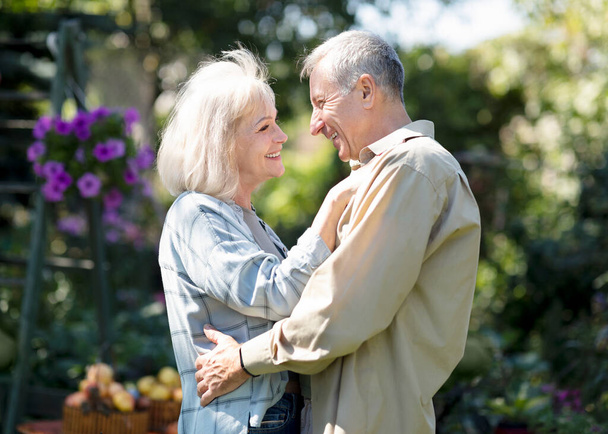 Счастливые старшие супруги танцуют в саду в теплый весенний день, смотрят друг на друга и улыбаются, отдыхают вместе на открытом воздухе - Фото, изображение