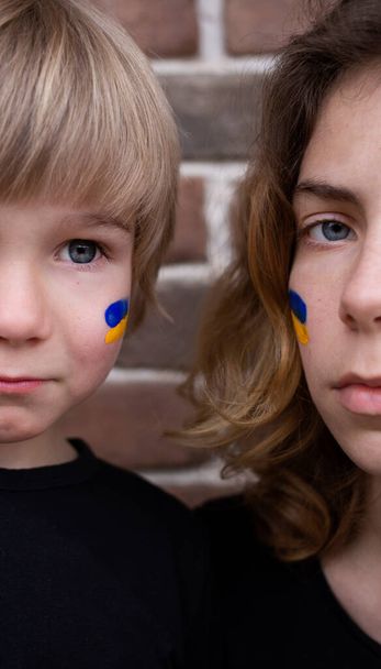 detailní záběr poloviny tváře smutného chlapce a mladé ženy s modrou a žlutou ukrajinskou vlajkou namalovanou na tvářích. Děti proti válce. stojan s Ukrajinou - Fotografie, Obrázek