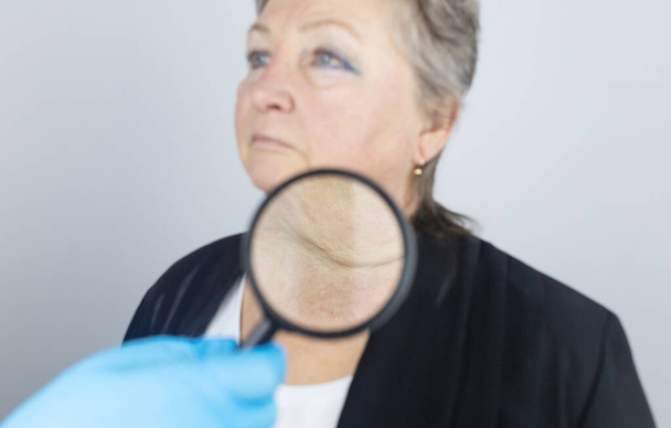 Starsza kobieta w recepcji chirurga plastycznego. Konsultacje w sprawie usuwania zmarszczek wieku na twarzy. Lekarz pokazuje, jakie procedury wykona. Mimiczne zmarszczki. Szkło powiększające w rękach - Zdjęcie, obraz