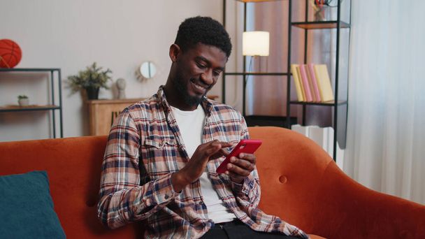 Радостный молодой человек, сидящий на диване с помощью смартфона, смотрит видео, делает покупки в Интернете, постукивает - Фото, изображение