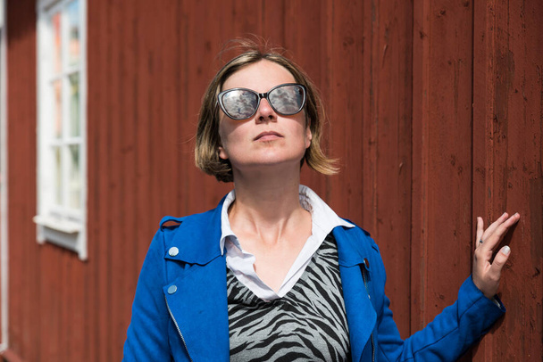 Oregrund, Uppland - Zweden - Dertigjarige vrouw met blauwe jas en zonnebril poserend voor de rode houten muur van een typisch Zweeds huis - Foto, afbeelding