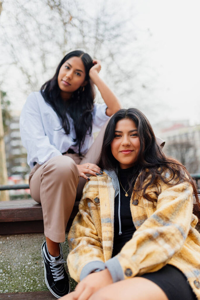 függőleges portré két fiatal spanyol lányról, akik egy városi parkban ülnek. Két latin nővér néz a kamerába.. - Fotó, kép
