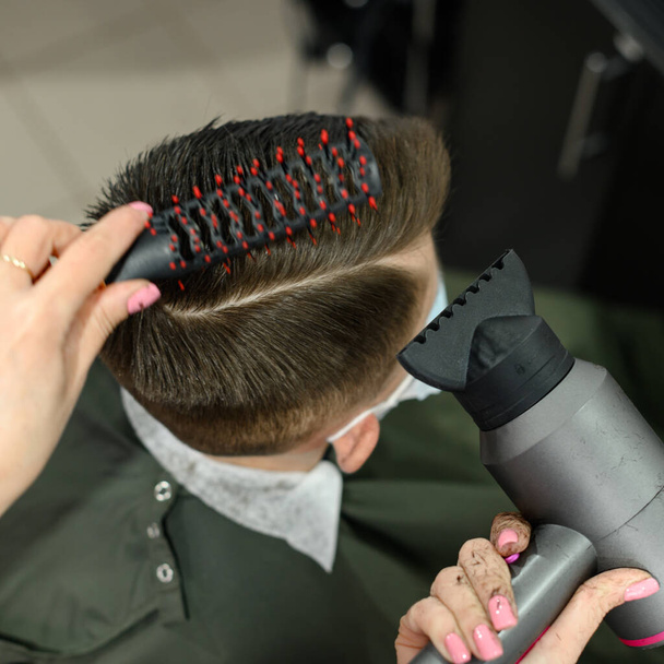Підліток отримує стрижку під час пандемії в перукарні, стрижку і сушку волосся після стрижки, укладання волосся після стрижки з фен
. - Фото, зображення