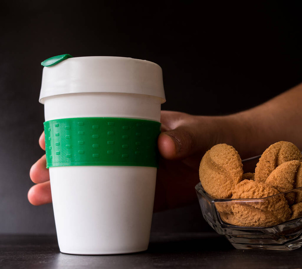 Biscuits au café et thermos en plastique pour boissons chaudes prises à la main sur fond noir - Photo, image