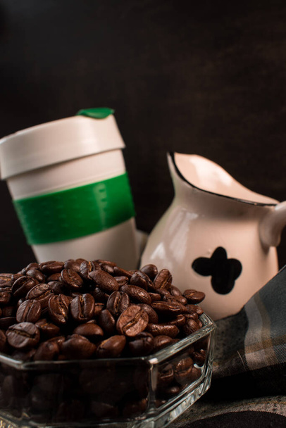 Кофейные зерна, молочный кувшин и пластиковый термос для горячих напитков, взятых рукой на черном фоне - Фото, изображение