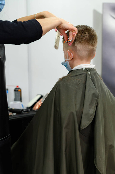 Kinderhaarschnitt im Friseursalon, stilvoller und moderner Haarschnitt für Schulkinder, Arbeit während der Pandemie, Kinderhaarschnitt mit der Schere. - Foto, Bild
