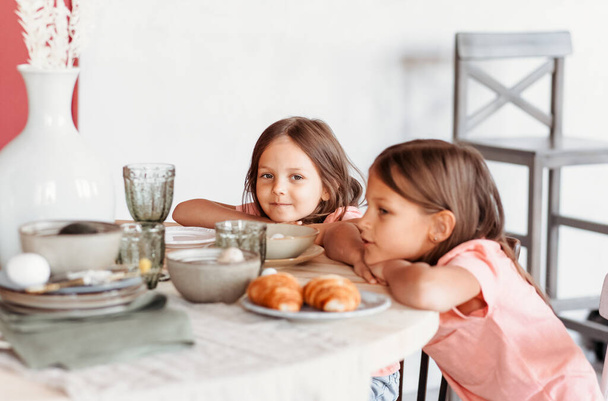 台所のテーブルに座ってる2人の少女。子供たちは家で朝食をとる。幸せな生活家族の瞬間. - 写真・画像