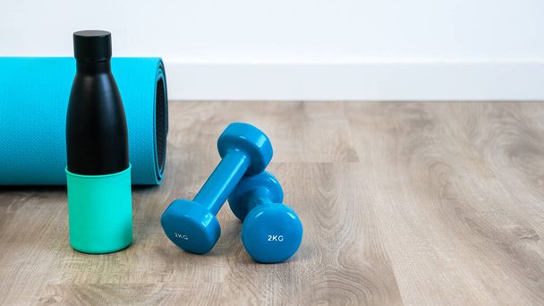 Ein Paar blaue Kurzhanteln und eine wiederverwendbare Wasserflasche aus Metall für Yoga auf dem Boden im Haus, gesunde Meditation am Morgen. Fitnessgeräte für Krafttraining. - Foto, Bild