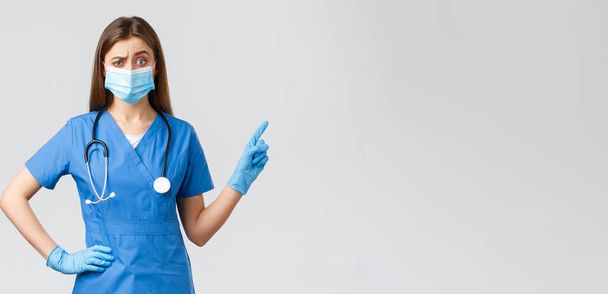Ковид-19, профилактика вируса, здоровье, медицинские работники и карантин. Скептически настроенная и недовольная медсестра или врач в синем халате, медицинская маска, указывающая пальцем направо, хмурящаяся сомнительная
 - Фото, изображение
