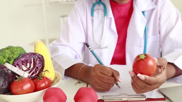 Professional nutrition doctor African-American female nutritionist sits down notes and benefits of tomatoes and broccoli for her hospital office patient meal plan. - Footage, Video