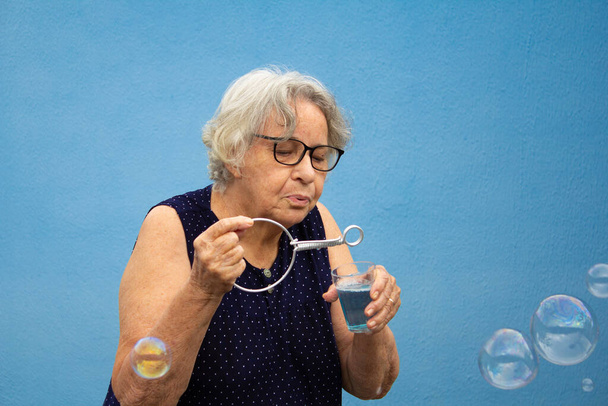 Пожилая женщина делает мыльные пузыри на синем фоне. - Фото, изображение