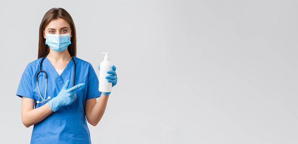 Covid-19, het voorkomen van virus, gezondheid, gezondheidswerkers en quarantaine concept. Jonge mooie vrouwelijke verpleegkundige of arts in blauwe scrubs, medisch masker en handschoenen, wijzend op de hand reiniger, zeep - Foto, afbeelding