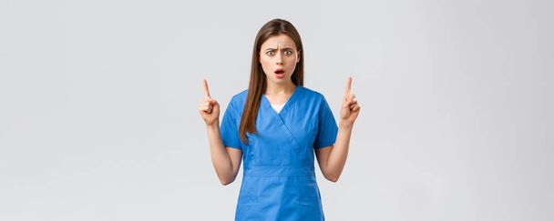 Healthcare workers, prevent virus, insurance and medicine concept. Oh gosh what this. Shocked and concerned nurse or doctor in blue scrubs, frowning upset, gasping pointing fingers up - Photo, Image