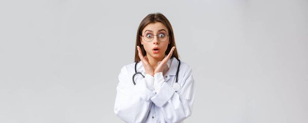 Healthcare workers, medicine, insurance and covid-19 pandemic concept. Surprised and impressed female doctor in glasses, white scrubs, gasping and staring astonished camera, grey background - Photo, Image