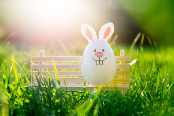 Creative Easter composition of Funny Easter egg with bunny ears sitting on a miniature bench on green grass background on sunny day. Bright festive mood. Fairy tale Easter. Selective focus. Copy space - Foto, imagen