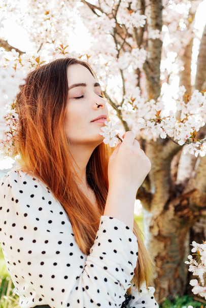Young brunette woman with closed eyes sitting near a pink blooming tree and enjoying the moment. Generation Z girl enjoy spring mood during a sunny day. Relax, freedom and personal fulfillment. - 写真・画像