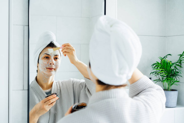 Woman in bathrobe with a towel on her head looking in the mirror and applying facial cosmetic clay mask on her face in bathroom. The concept of cosmetic procedures for skin care. Home Beauty self-care - Fotografie, Obrázek