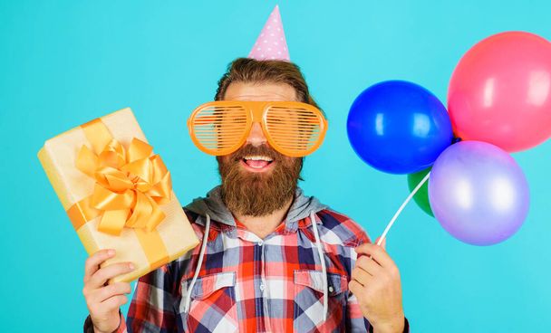 Ευτυχισμένος άνθρωπος γιορτάζει τα γενέθλια. Γενειοφόρος με καπέλο διακοπών με μπαλόνια και κουτί δώρου. Ώρα για πάρτι. - Φωτογραφία, εικόνα