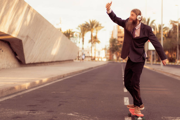 Hipster homem de negócios usar um fone de ouvido sem fio e skate smartphone perto de escritório de negócios. Conceito de transporte. - Foto, Imagem