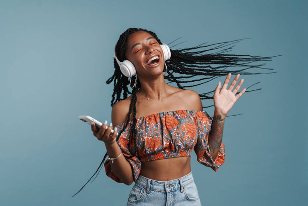 Young black woman singing while listening music with headphones isolated over blue background - Фото, зображення