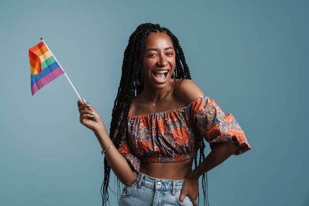 Giovane donna nera ridere mentre posa con bandiera arcobaleno isolato su sfondo blu - Foto, immagini