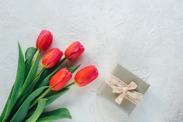 Prezent w pudełku i czerwone tulipany na białym, teksturowanym tle. Koncepcja wiosennych wakacji. Widok z góry, płaski, przestrzeń do kopiowania - Zdjęcie, obraz