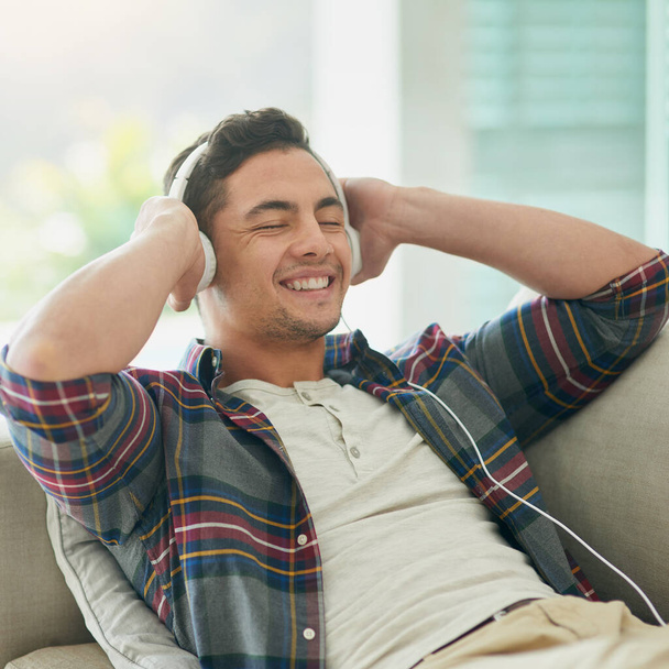 Сделай неделю потише, включи музыку. Фотография молодого человека, отдыхающего на диване и слушающего музыку в наушниках дома. - Фото, изображение
