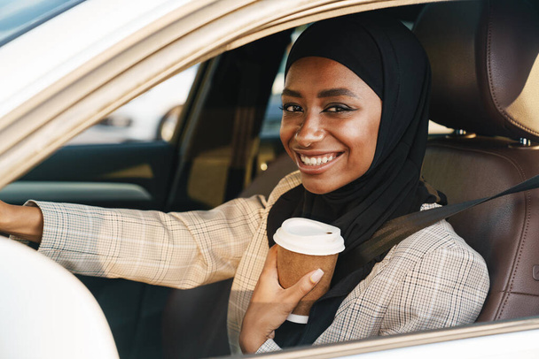 Black muslim woman wearing headscarf drinking coffee while driving car - Fotoğraf, Görsel