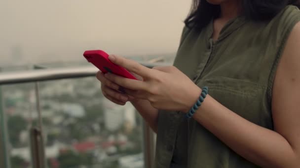 Primeros planos de las mujeres usando teléfonos inteligentes revisando las redes sociales y charlando con amigos mientras están de pie junto a un balcón en una azotea en el condominio durante el atardecer. - Imágenes, Vídeo