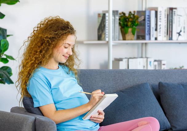 Caucasian happy beautiful long curly hair healthy pregnancy mother in casual outfit sitting smiling holding pencil writing taking note on diary notebook planning for newborn childbirth in living room. - Foto, immagini