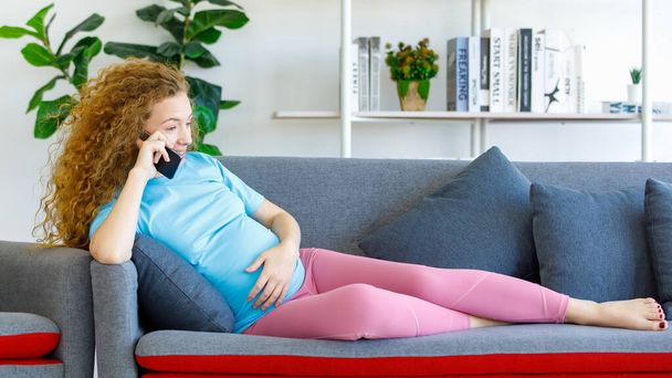 Caucasian happy beautiful long curly hair healthy pregnancy mother in casual outfit sitting laying down on cozy sofa touching tummy belly having conversation on call with smartphone in living room. - Фото, зображення
