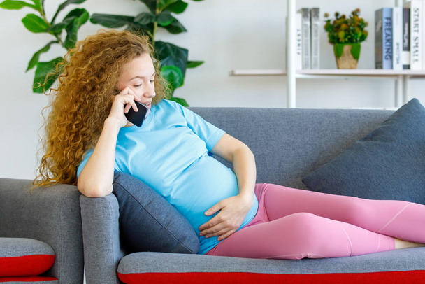 Caucasian happy beautiful long curly hair healthy pregnancy mother in casual outfit sitting laying down on cozy sofa touching tummy belly having conversation on call with smartphone in living room. - Photo, Image